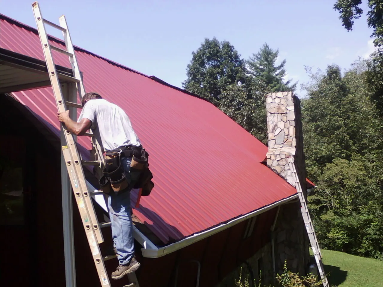 Roofer on a ladder beside a red metal roof and stone chimney — Soffit & Fascia Repair by Roofing Brothers in Brunson